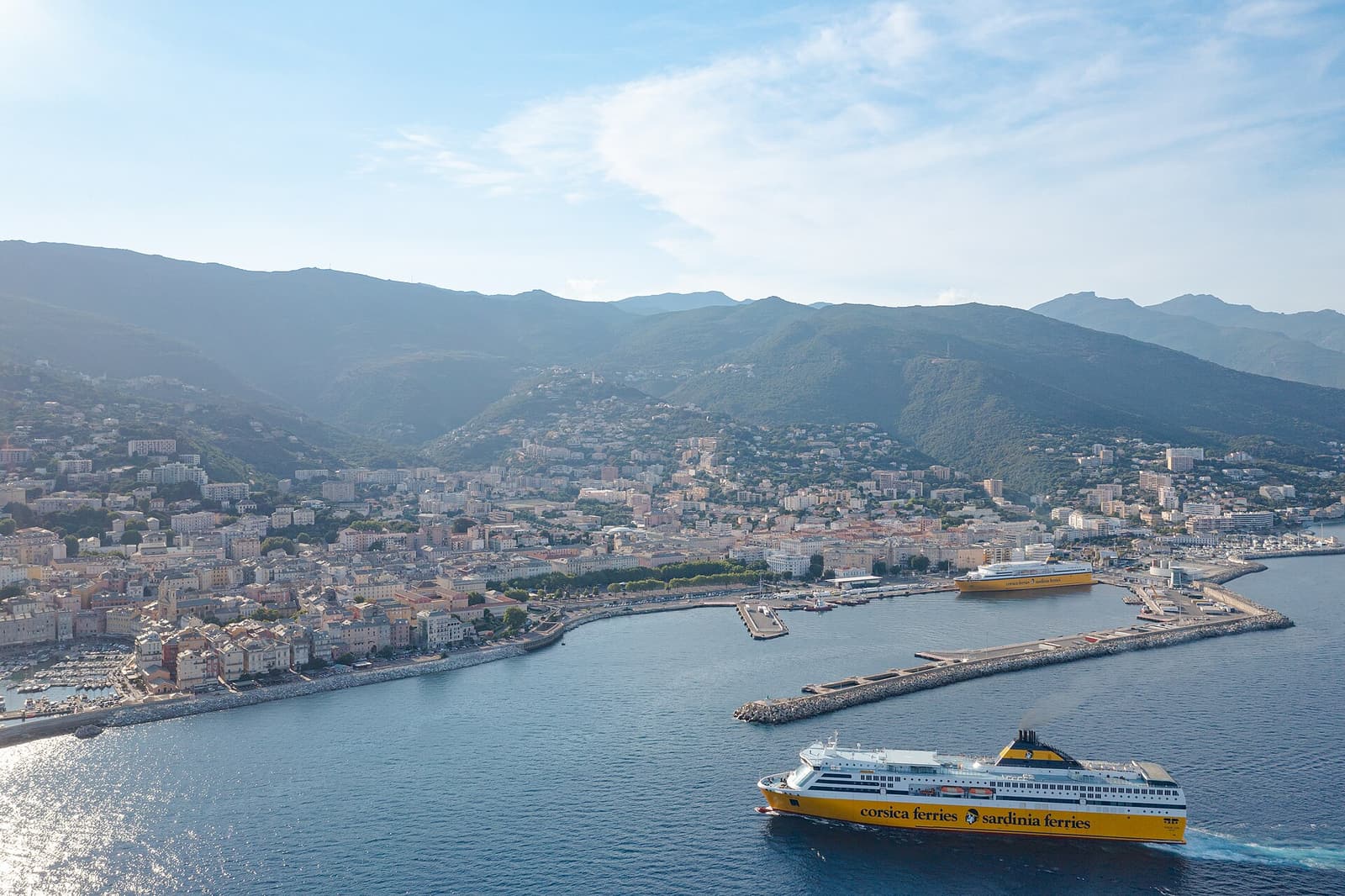 A yellow ferry leaving the port of Bastia with the town and mountains behind