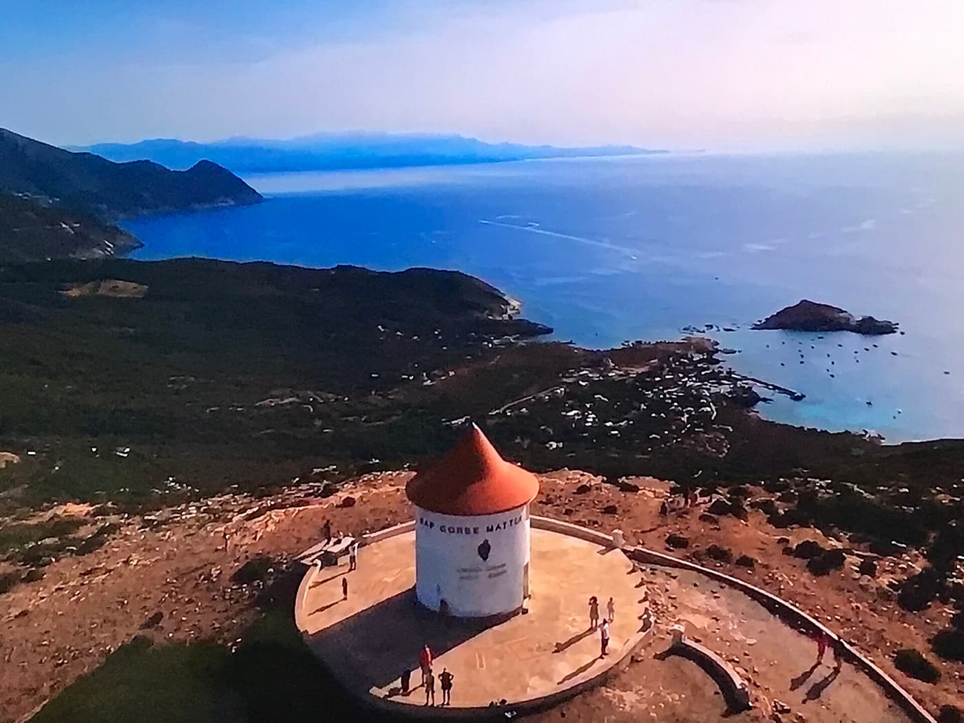 Aerial view of the white, red-roofed Moulin Mattei on its hilltop, with the north-west coast of Cap Corse and the islet of Centuri below